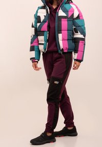 Kleurrijke gewatteerde jas met turquoise, roze, zwarte en witte geometrische patronen. In combinatie met bordeauxrode broek en zwarte sportschoenen.