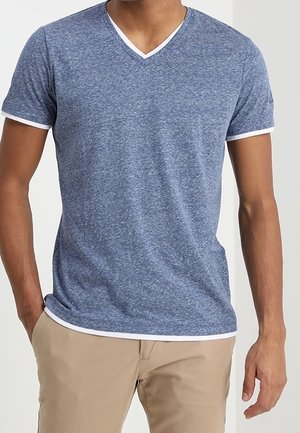 Homme portant un T-shirt bleu à manches courtes et col en V avec une bordure blanche, associé à un pantalon beige, debout avec une main dans la poche.
