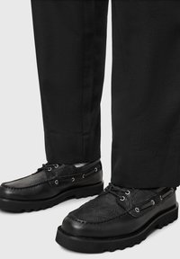 Zapatos negros con paneles texturizados, diseño con cordones y ojales de metal. Suela gruesa con dibujo; combinados con pantalones negros.