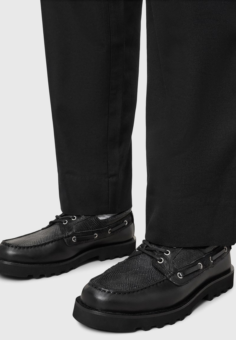 Zapatos negros con paneles texturizados, diseño con cordones y ojales de metal. Suela gruesa con dibujo; combinados con pantalones negros.