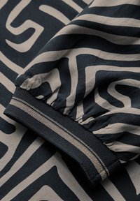 Tessuto nero e beige con un design a motivo ondulato. Il polsino elastico presenta strisce a contrasto sul bordo. Texture liscia, aspetto leggero.