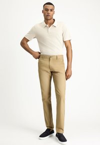 Light beige polo shirt with short sleeves, paired with tan trousers and dark blue slip-on shoes. Smooth fabric, tailored fit, clean lines.