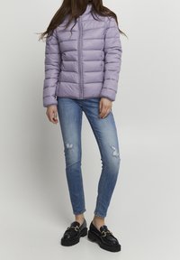 Lawendowa pikowana kurtka puchowa z wysokim kołnierzem i zamkiem z przodu. Dopasowana do podniszczonych niebieskich dżinsów i czarnych mokasynów.