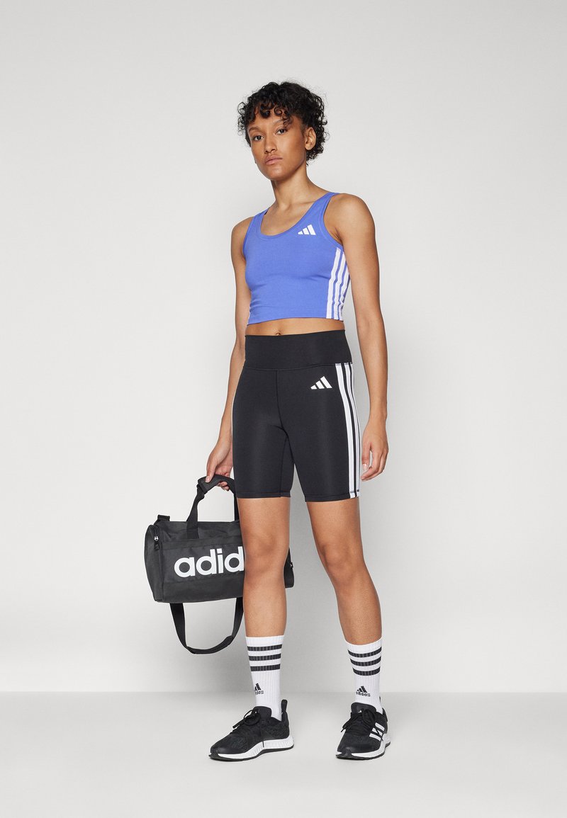 Top corto azul claro con rayas blancas, pantalones cortos negros de cintura alta con detalles blancos, zapatillas negras y una bolsa negra con el logo de Adidas.