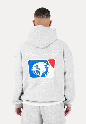 Mand iført lysegrå hættetrøje og matchende joggingbukser, set bagfra, hættetrøjen viser blå, hvid og rød brølende katte-logo med tekst over.