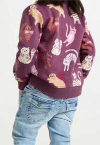 Cranberry-kleurige sweatshirt met speelse kattenpatronen in verschillende kleuren, gecombineerd met lichtblauwe jeans met twee achterzakken en decoratieve stiksels.