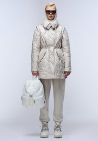Parka imbottita beige con colletto alto, vita regolabile con cordino e dettagli con tasche. Abbinata a pantaloni jogger beige chiaro e uno zaino bianco trapuntato.