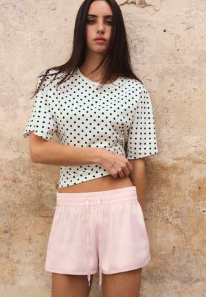 Femme aux longs cheveux foncés portant un haut blanc à pois et un short rose clair, debout devant un mur beige texturé.