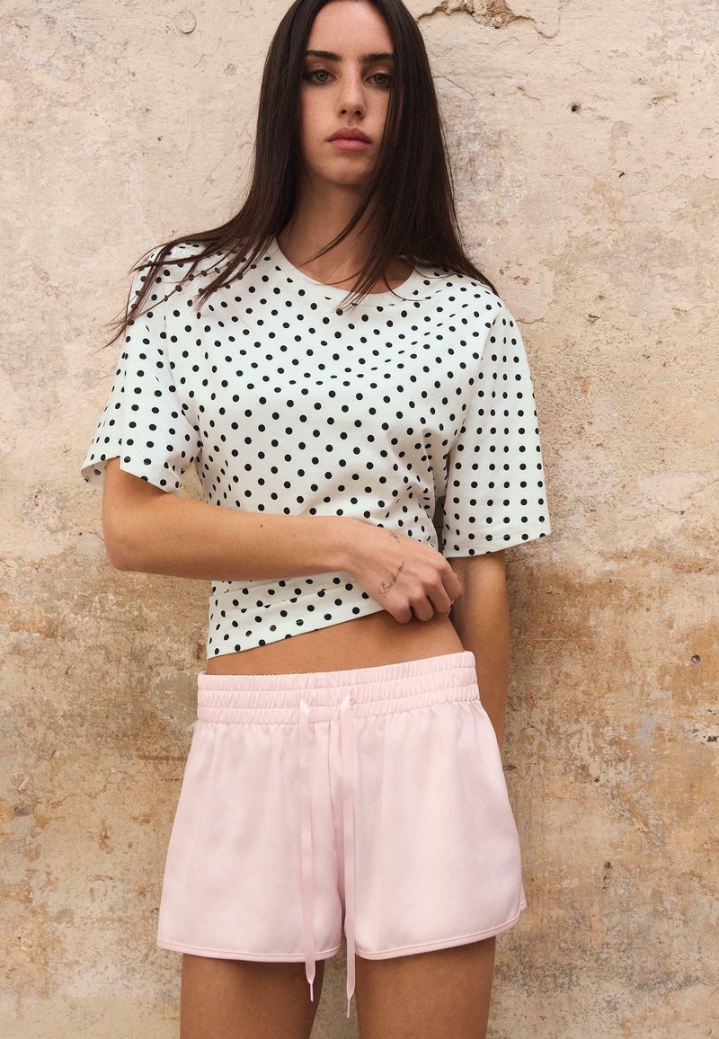 Femme aux longs cheveux foncés portant un haut blanc à pois et un short rose clair, debout devant un mur beige texturé.