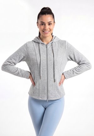 Jonge vrouw die glimlacht, draagt een lichtgrijze hoodie met rits en lichtblauwe legging, staande met handen in de zij tegen een witte achtergrond.