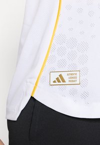 T-shirt en maille blanche avec des bordures jaunes, présentant un motif texturé et un badge doré indiquant "PRODUIT LICENCIÉ AUTHENTIQUE" avec un logo.