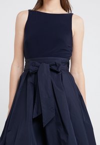 Marinblå klänning med en figurnära överdel och breda axelremmar, med en plisserad kjol och en stor rosett i midjan, tillverkad av ett slätt material.