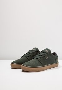 Tenisi negri din suede cu accent verde, având o talpă din cauciuc și șireturi plate. Logo-ul este vizibil pe lateral și pe limbă.
