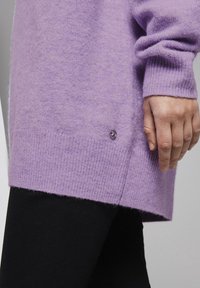 Pull en tricot violet avec des poignets côtelés, un ourlet arrière plus long et un petit détail de bouton décoratif sur le côté. Associé à des leggings noirs.