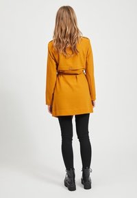 Musztardowożółty płaszcz z paskiem, długimi rękawami i klapą z tyłu, w połączeniu z czarnymi obcisłymi jeansami i masywnymi czarnymi botkami.