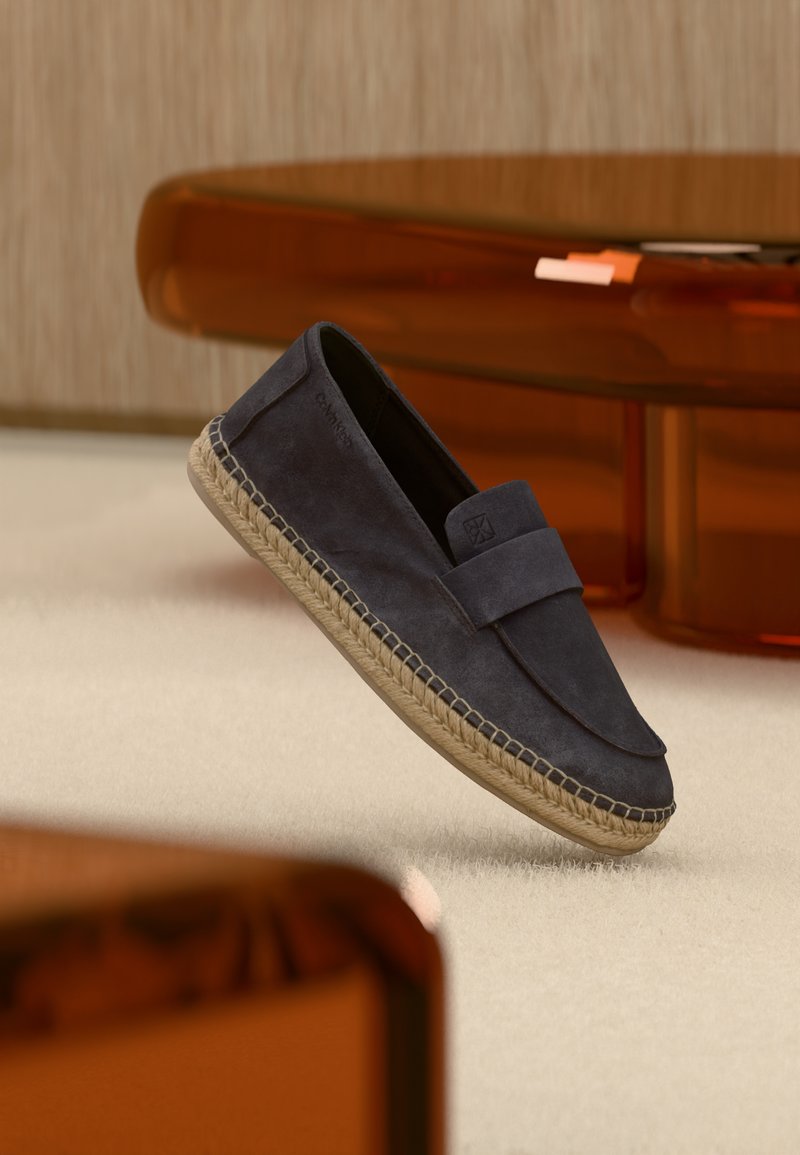 Mocassin en daim bleu marine avec semelle en jute tissé flottant au-dessus d'un tapis beige, avec un mobilier ambre translucide en arrière-plan.