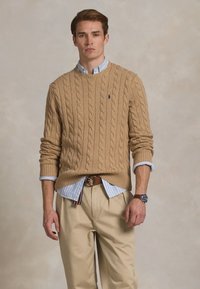 Jonge man in een beige kabeltrui over een blauw gestreept shirt, beige broek en een horloge, staand tegen een neutrale achtergrond.