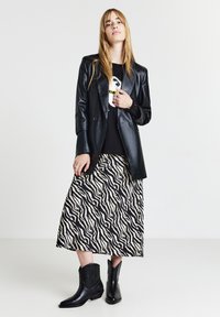 Giovane donna che indossa una giacca di pelle nera, una t-shirt con grafica, una gonna midi a strisce zebrate e stivaletti neri, in piedi contro uno sfondo bianco.