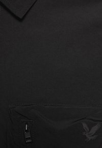 Μαύρο ύφασμα με τσέπη με φερμουάρ και ένα σκούρο γκρι λογότυπο αετού τοποθετημένο στο κάτω δεξί μέρος της τσέπης.