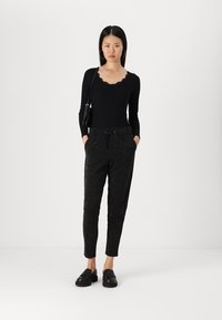 Haut noir à manches longues avec un décolleté en forme de vague, associé à un pantalon sombre pailleté et des chaussures noires, avec les mains dans les poches et un petit sac.