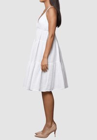 Vestido de lino blanco con dobladillo acampanado, tirantes finos y diseño en capas. Combinado con zapatos de tacón nude.