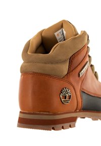 Bota de cuero marrón con un cuello de ante color marrón claro, presentando un panel texturizado negro, acento de logo metálico y una suela resistente con tacos de tracción.