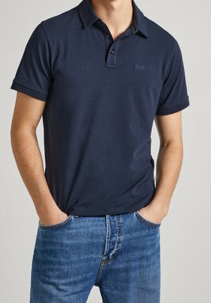 Homme portant un polo bleu marine à manches courtes, légèrement rentré dans un jean en denim bleu, mains dans les poches, sur fond uni.