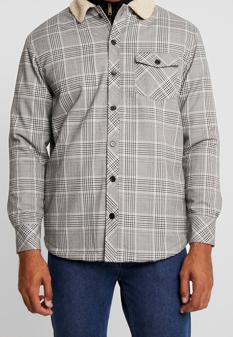 Homme portant une chemise à carreaux grise boutonnée avec une poche poitrine et un col sherpa beige, associée à un jean bleu sur un fond uni.