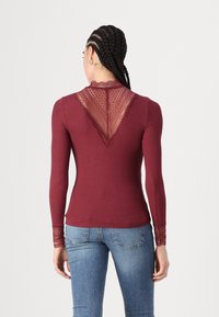 Burgundyröd långärmad topp med spetsbakpanel, ribbad textur och spetsdetaljer vid ärmsluten, tillsammans med blå jeans.