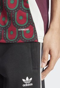 Camisa de manga corta color marrón y con estampado de diseño en verde y burdeos, y pantalones de chándal negros que exhiben el logo y los acentos blancos de Adidas.