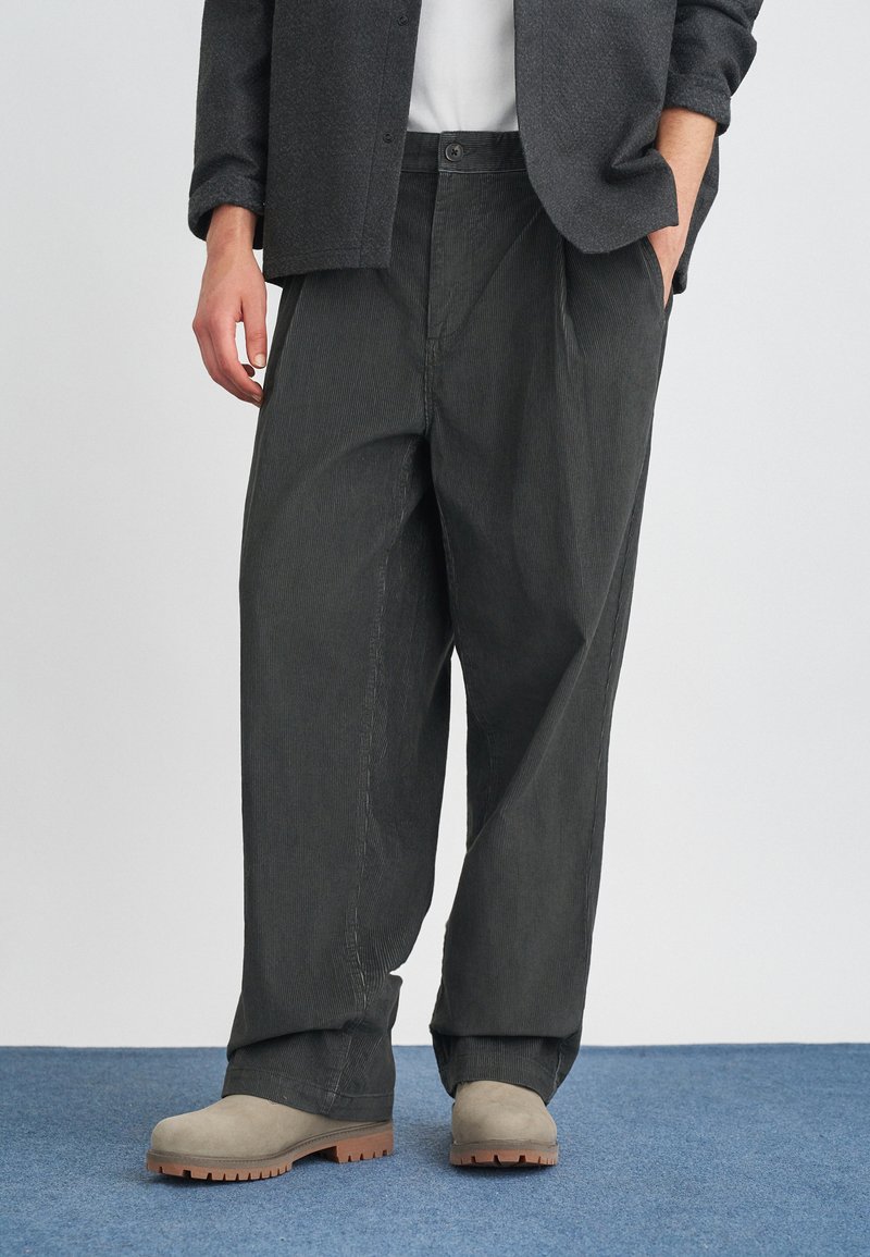 Pilkos korduroy kelnės su laisvu kirpimu, su šoninėmis kišenėmis ir mygtuku liemens srityje, derinamos su pilkais zomšiniais batais.