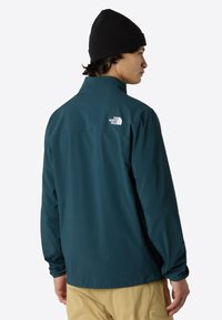 Teal waterproof jacket with a high collar, long sleeves, and a white logo on the back. Worn with beige pants and a black beanie.