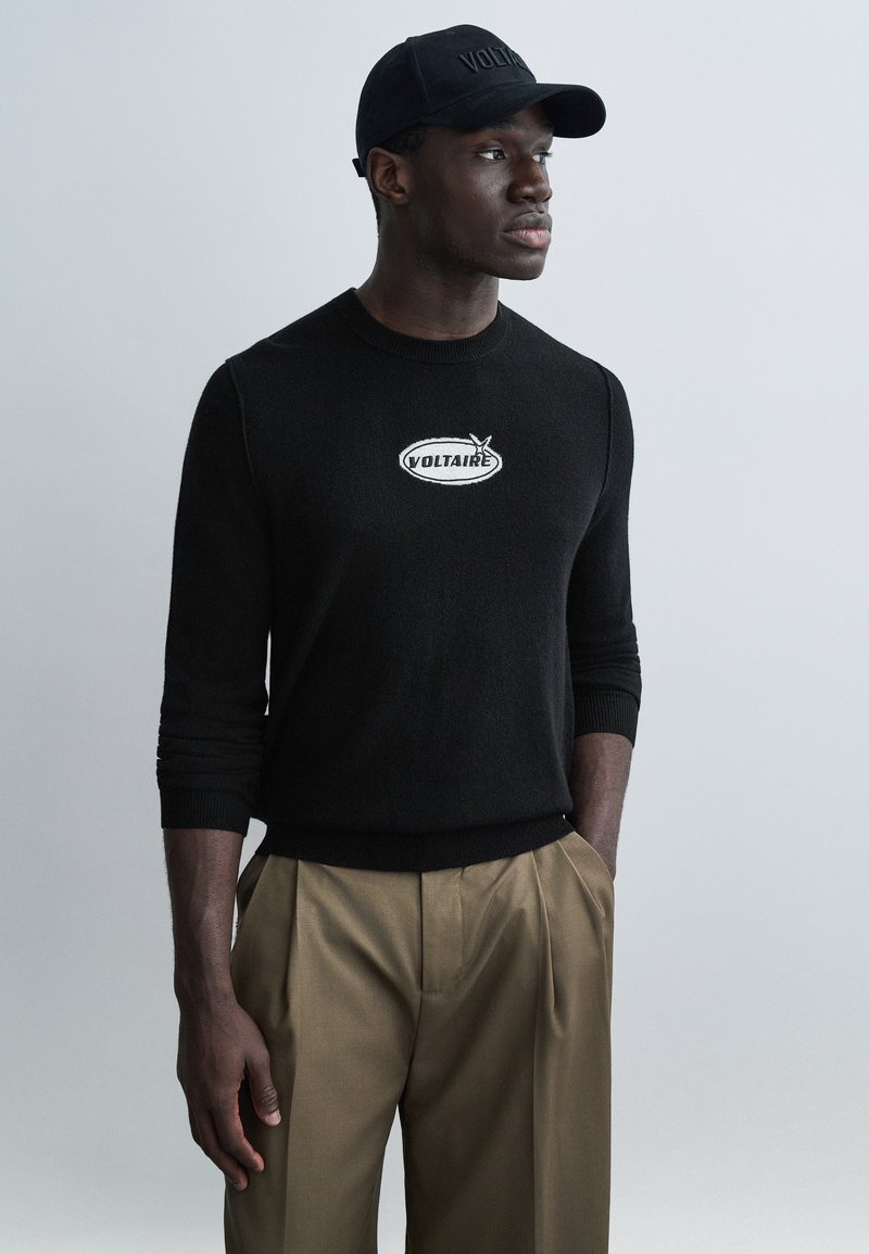 Black knit sweater with "VOLTAIRE" embroidered in white, paired with tan pleated trousers and a black cap featuring the same logo.