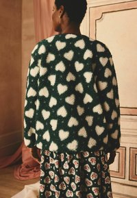 Pullover verde pelo con motivo a cuori bianchi, abbinato a una gonna scura a fiori con motivi a forma di cuore e orlo arricciato.