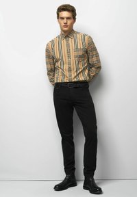 Jonge man die tegen een muur staat, gekleed in een beige, rode en zwarte gestreepte overhemdd, zwarte broek, gevlochten riem en zwarte laarzen.