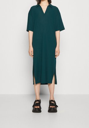 Tyrkysově modré šaty po kolena s výstřihem do V a krátkými rukávy. Mají boční rozparky a texturovanou látku. Kombinovány s černou obuví.