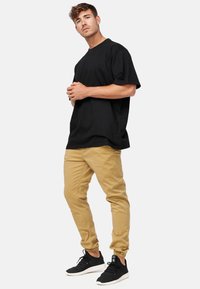 Młody mężczyzna stoi z złączonymi rękami, ubrany w luźną czarną koszulkę, beżowe spodnie dresowe i czarne trampki na białym tle.