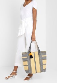 Borsa tote gialla e nera con motivo a fantasia, realizzata in tessuto testurizzato, presenta un accento con nappina e due manici robusti, forma rettangolare e apertura superiore.