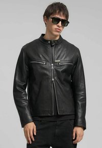 Zwarte leren motorjack met een opstaande kraag, ritssluiting aan de voorkant en twee borstzakken met rits. Soepele textuur en nauwsluitend ontwerp.