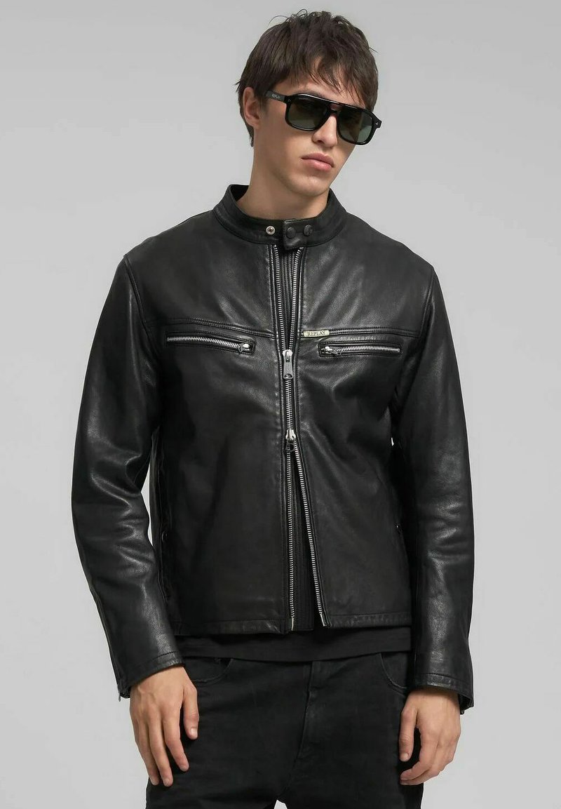 Zwarte leren motorjack met een opstaande kraag, ritssluiting aan de voorkant en twee borstzakken met rits. Soepele textuur en nauwsluitend ontwerp.