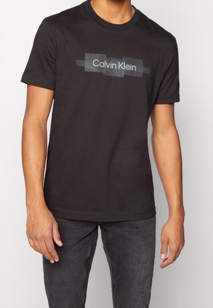 Mężczyzna w czarnym t-shircie Calvin Klein i ciemnoszarych dżinsach, stojący z rękami swobodnie opuszczonymi wzdłuż ciała na jednolitym tle.
