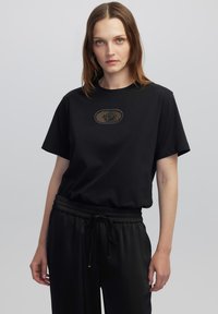 Camiseta negra de manga corta con cuello redondo y un parche ovalado texturizado en el pecho, combinada con pantalones negros de cintura elástica.