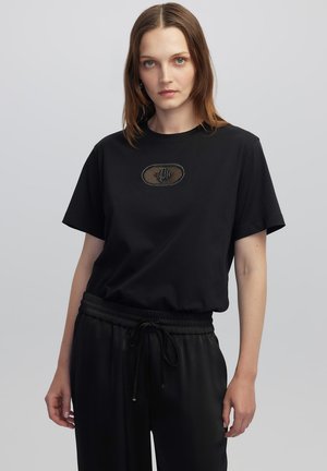 Camiseta negra de manga corta con cuello redondo y un parche ovalado texturizado en el pecho, combinada con pantalones negros de cintura elástica.