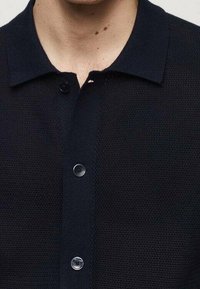 Černý pletený polo triko s knoflíkovou légou a texturovanou tkaninou. Má klasický límec a hladké tmavé knoflíky.