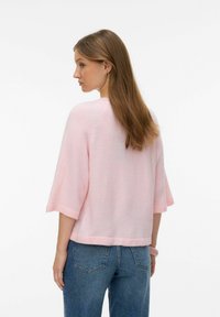 Svetlo roza prevelik pulover s tremi četrtinami rokavov, mehke teksture in zaokroženim robom. Kombiniran z visokim pasom modrih kavbojk.