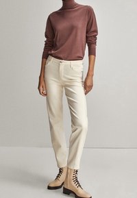 Kermansinväriset housut yhdistettynä ruosteenväriseen mock-turtleneck-neuleeseen; mallilla on myös päällään beigejä nauhakengät, joissa on massiivinen pohja.