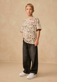 Beige grafisk t-shirt med Marvel-designs, parret med løse mørkegrå denimjeans og hvide sneakers med sorte accenter.
