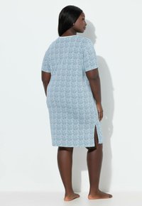 Mujer con cabello largo y oscuro, vistiendo un vestido azul claro con estampado, de largo hasta las rodillas, con mangas cortas y una abertura en el costado, de pie descalza mirando una pared blanca.