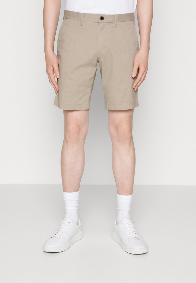 Homme portant un short beige au genou, des chaussettes blanches et des baskets blanches, debout devant un fond clair uni.