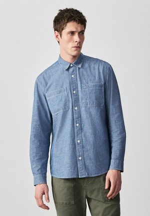 Mies, joka pukeutunut pitkähihaiseen siniseen chambray-paitaan, jossa on kaksi rintataskua, ja oliivinvihreisiin housuihin, seisoo yksinkertaista vaaleaa taustaa vasten.